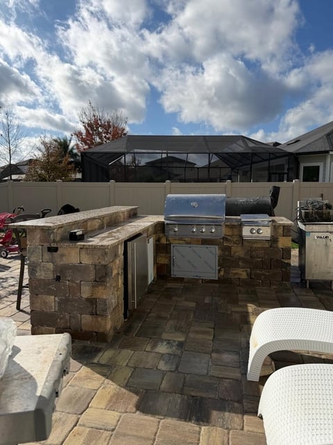 Outdoor kitchen with built-in stone grill and countertops on patio, under blue sky with white house in background