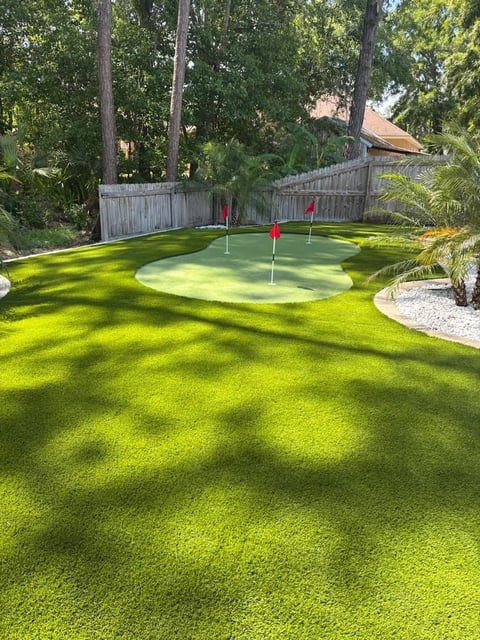 Backyard putting green with two holes marked by red flags, surrounded by lush green grass and tall trees, with a wooden fence and house in background
