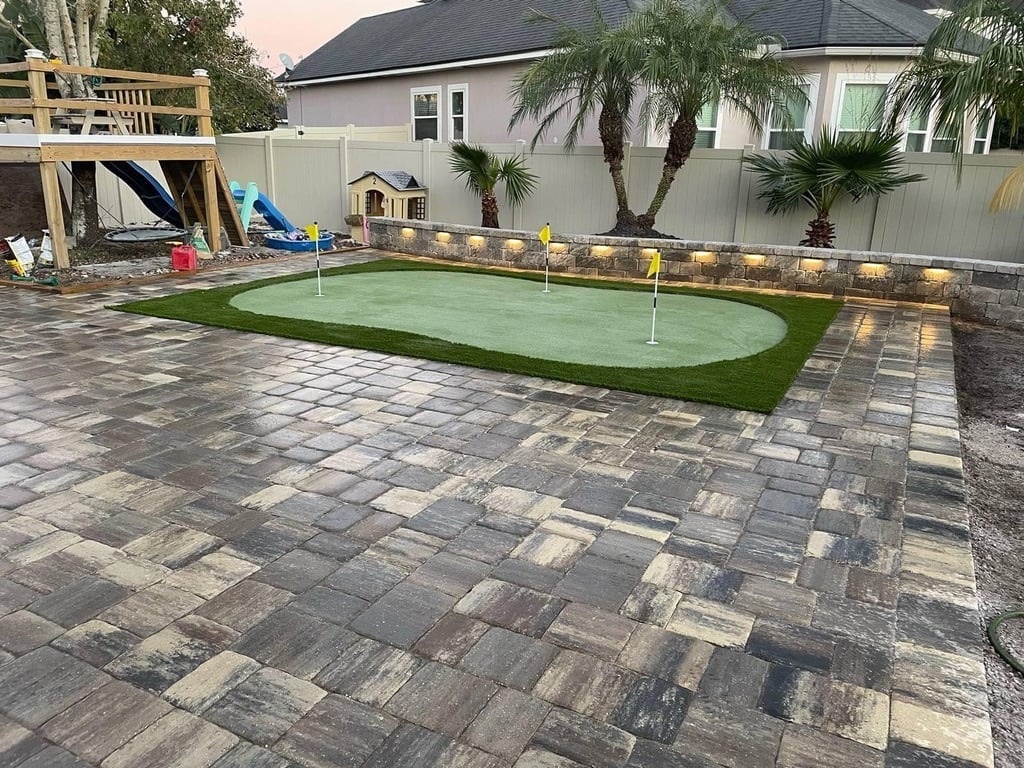 Backyard patio with paver stone flooring, artificial putting green, and white house with palm trees in background