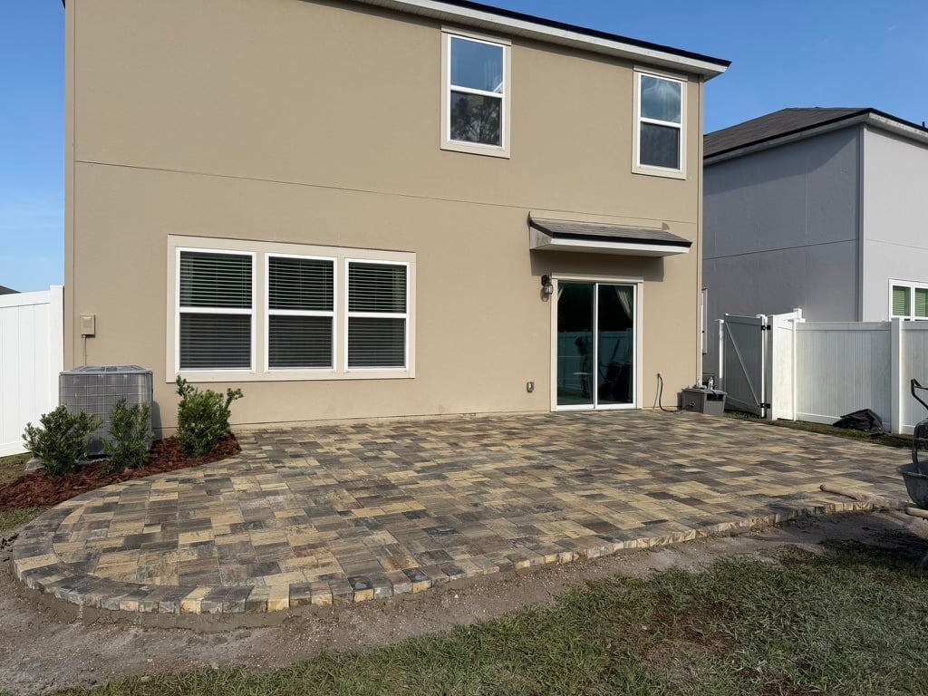 Modern two-story beige house with paver patio, large windows, and dark entry door on sunny day