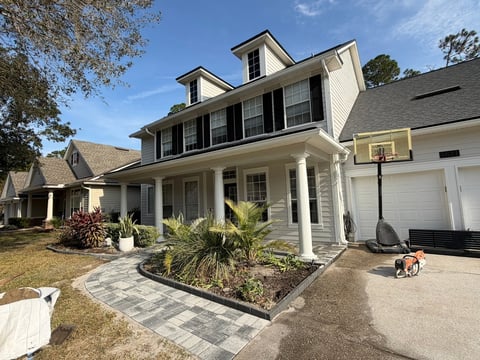 Two-story white house with black shutters, front porch, columns, and attached garage on a sunny day with landscaping