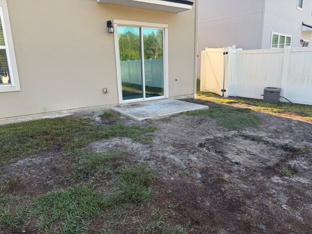 Backyard patio area with patchy grass, sliding glass doors, white vinyl fence, and construction materials on right side.