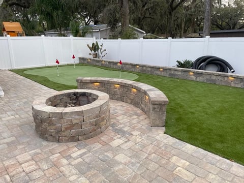 Backyard with circular stone fire pit, patio, artificial putting green with flags, and white fence.